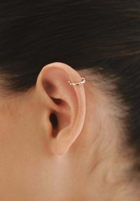 Pendiente de aro dorado llevado en la parte superior de la oreja, con una textura torcida y un acabado suave. Primer plano de la oreja y los detalles del pendiente.