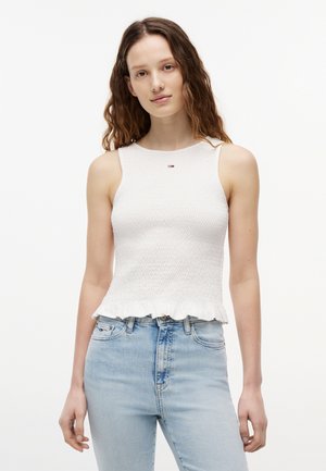 Młoda kobieta o falowanych włosach, ubrana w białą, bezrękawnikową teksturowaną bluzkę i jasnoniebieskie dżinsy, stojąca na tle jednolitego białego tła.