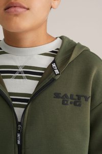 Olivovo zelená mikina na zips s čiernym logom, skombinovaná s prúžkovanou svetlosivou a zelenou mikinou s okrúhlym výstrihom. Obsahuje textúrovanú tkaninu a rebrované manžety.