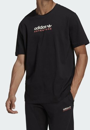 Homme portant un t-shirt noir Adidas Adventure et un pantalon noir assorti avec logo et texte sur la poitrine et la cuisse gauche.