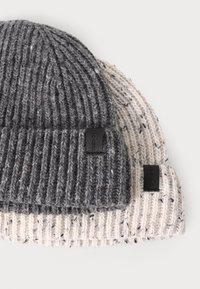 Dos gorros de punto: uno de color gris oscuro y otro crema con manchas. Ambos presentan una textura de canalé y un pequeño parche de logo negro.