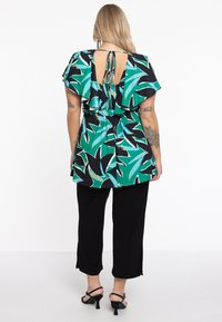 Kurzärmlige, grün-schwarze Bluse mit floralem Muster und geschichteten Rüschendetails auf der Rückseite, kombiniert mit schwarzen, cropped Hosen und schwarzen, Absatzsandalen.