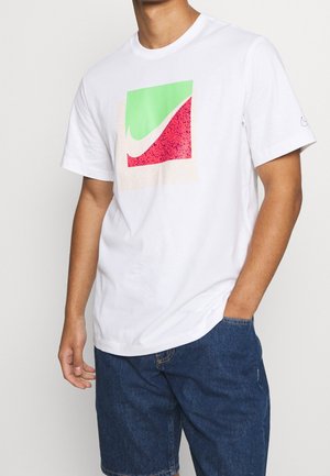 Hombre con camiseta blanca que lleva un logotipo cuadrado colorido y pantalones cortos de denim azules, con una mano en el bolsillo frente a un fondo liso.