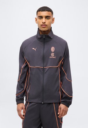 Junger Mann trägt eine schwarze Puma AC Milan Fußball-Zipjacke mit orangenen Paspeln, steht vor einem einfarbigen hellen Hintergrund.
