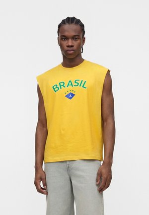 Man met gevlochten haar draagt een geel mouwloos shirt met de tekst "BRASIL" en sterren, gecombineerd met lichtgrijze broek, staand tegen een witte achtergrond.