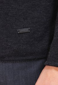 Svart texturerad tyg med en liten rektangulär metallskylt som står "BOSS ORANGE" nära en persons handled och mörk denim nedanför.