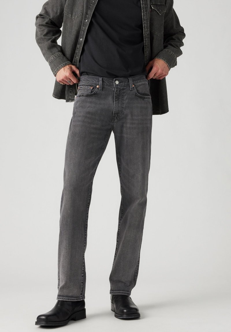 Šedé džíny z denimu s rovnými nohavicemi, pět kapsami a kontrastním prošíváním, spárované s černými koženými botami a tmavým tričkem.