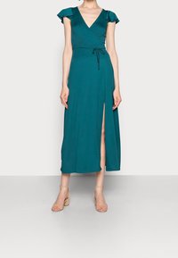 Tee-farbenes Wickelkleid mit kurzen, rüschierten Ärmeln, tiefem V-Ausschnitt, seitlichem Schlitz und verstellbarem Taillengürtel. Mit neutralen Blockabsatzsandalen kombiniert.