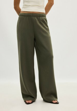 Person, die lockere, weite olivgrüne Hosen und schwarze Sandalen mit offenen Zehen trägt, von der Taille abwärts vor einem einfarbigen Hintergrund sichtbar.