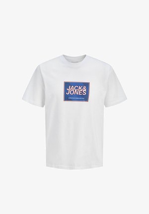 Garçon blond debout, portant un t-shirt blanc Jack & Jones, un jean gris ample et des baskets grises, regardant vers la gauche sur un fond uni.