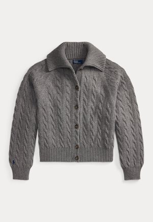 Femme portant des lunettes de soleil noires, un cardigan gris à torsades, un pantalon large gris, des chaussures noires, avec un sac à épaule noir, debout devant un fond uni.