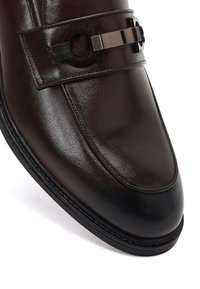 Mocassino in pelle marrone scuro con punta nera, suola nera e dettaglio in metallo sulla fascia superiore.