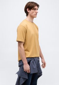 Νεαρός άνδρας με καστανά μαλλιά, που φοράει μουσταρδί κίτρινη μπλούζα και μπλε καρό πουκάμισο δεμένο γύρω από τη μέση, κοιτάζει προς τα δεξιά σε απλό φόντο.