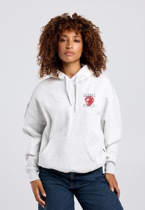 Vrouw met krullend haar draagt een lichtgrijze hoodie met een kleine rode opdruk en een jeans, staand tegen een effen lichte achtergrond.