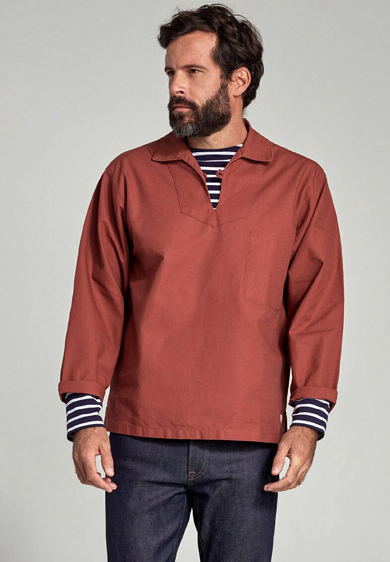 Camicia rustica a maniche lunghe con scollo a V e taschino sul petto, indossata sopra una maglietta a maniche lunghe a righe blu e bianco, abbinata a jeans scuri.