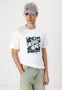 Camiseta blanca con un estampado oscuro de flores y texto en negrita. Gorra gris con varios parches coloridos. Vaqueros azul claro con un corte relajado.