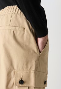 Beige cargo bukser i et glat, tekstureret stof med et ternet mønster, elastisk talje, sidelomme og sorte knappedetaljer.