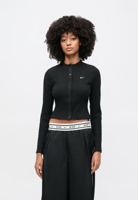 Μαύρη ριμπ ζακέτα με στρογγυλή λαιμόκοψη και κούμπωμα με κουμπιά, με μικρό λευκό λογότυπο Nike στο αριστερό στήθος. Μακριά μανίκια, cropped σχεδιασμός.