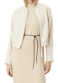 Giacca bomber beige chiaro con texture liscia, chiusura con zip e polsini a coste, indossata sopra un vestito beige a pieghe con una cintura nera.