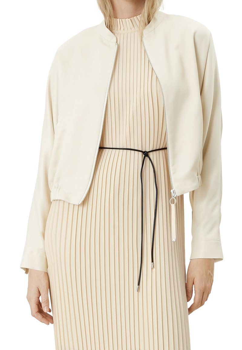 Giacca bomber beige chiaro con texture liscia, chiusura con zip e polsini a coste, indossata sopra un vestito beige a pieghe con una cintura nera.