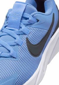 Svijetloplava atletska cipela s teksturiranim materijalom, crnim swoosh logotom, bijelim potplatom i zvjezdastim logom na jeziku. Ima vezice i ojačana područja.