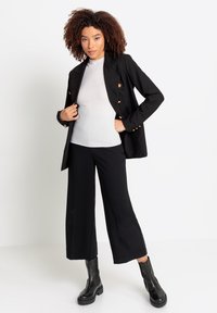 Zwarte blazer met gouden knoopaccenten, gecombineerd met een geribbelde witte top en zwarte wijde broek, aangevuld met zwarte enkellaarsjes.