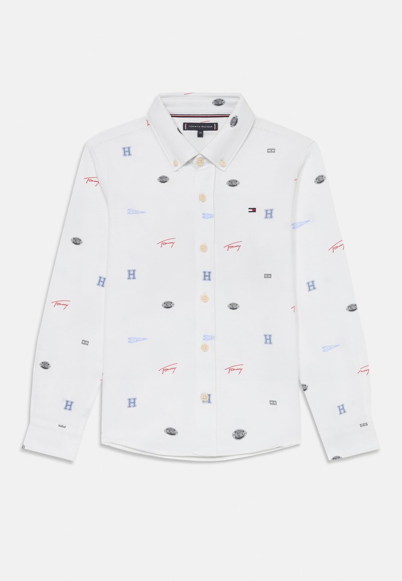 Bela bombažna srajca z dolgimi rokavi, ki ima različne logotipe v rdeči, modri in črni barvi. Ovratnik s trakovi z bež gumbi. Prilagojen kroj.