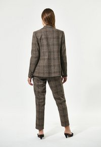 Completo su misura a quadri marroni con giacca aderente e pantaloni affusolati. La texture è liscia con un leggero lucido, accentuata da mule a tacco nero.