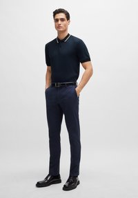 Camisa polo azul marino con cuello y detalles en dorado, combinada con pantalones chinos ajustados azul marino y botas negras con cordones. Diseño simple y moderno.