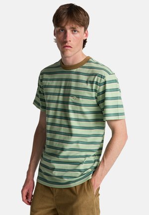 Młody mężczyzna o jasnobrązowych włosach, ubrany w zielono-beżową koszulkę w paski i brązowe spodnie, stoi z jedną ręką w kieszeni.