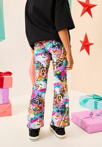 Lapsi, jolla on päällään värikkäät jouluteemaiset housut, joissa on porojen ja lumiukkojen kuvasto, seisoo lahjapakkausten ja punaisten tähden koristeiden keskellä.