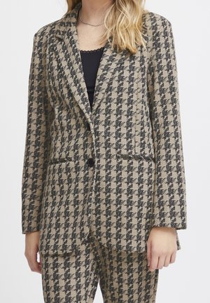 Kobieta ubrana w beżowo-czarną marynarkę w jodełkę i dopasowane spodnie, czarną bluzkę oraz srebrny łańcuszek na szyi.