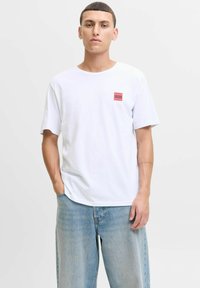 Mann in weißem Jack & Jones T-Shirt und lockeren hellblauen Jeans, steht mit einer Hand in der Tasche vor einem schlichten Hintergrund.