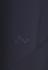 Tamno plava aktivna odjeća s rebrastom teksturom, ravnim šavovima i malim okruglim logotipom u različitim nijansama plave.