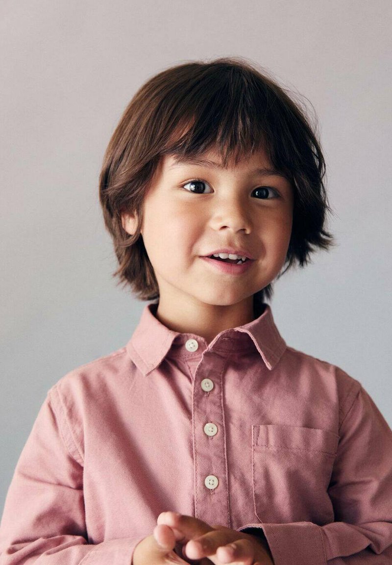 Malé dítě v světlé růžové košili na knoflíky s límečkem a přední kapsou. Vlasy jsou středně dlouhé a tmavě hnědé. Světlé pozadí.