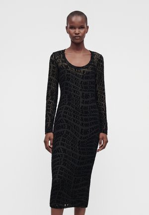 Wysoka osoba ubrana w czarną, przezroczystą sukienkę z długim rękawem, zdobioną powtarzającym się napisem „VERSACE JEANS COUTURE”.