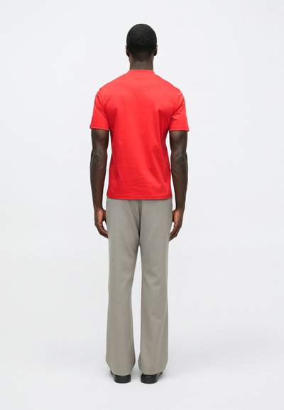 Άνδρας που στέκεται με την πλάτη στραμμένη, φορώντας ένα κόκκινο κοντομάνικο πουκάμισο, ανοιχτό γκρι παντελόνι και μαύρα παπούτσια, μπροστά σε λευκό φόντο.