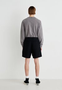 Graues Langarmshirt und schwarze Shorts mit lockerer Passform. Schwarze Schnürschuhe mit weißen Sohlen. Schlichter Hintergrund, minimalistischer Stil.