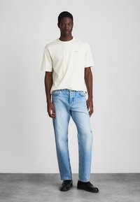 Laki-laki berdiri mengenakan kaos putih lengan pendek, jeans biru muda, dan sepatu hitam di latar belakang abu-abu polos.