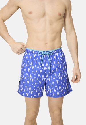 Hombre con pantalones cortos de baño azules con patrones de velero blanco y cordón de color azul claro, de pie contra un fondo claro y sencillo.