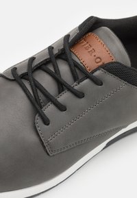 Basket grise en matériau synthétique lisse, avec des lacets noirs, un accent en cuir beige et une semelle en caoutchouc blanc avec un détail latéral texturé.