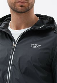 Sort hættetrøje med lynlås foran, der har hvid tekst "DIN ENESTE GRÆNSE ER DIN HJERNES GRÆNSE," og tekstureret indvendig foring.