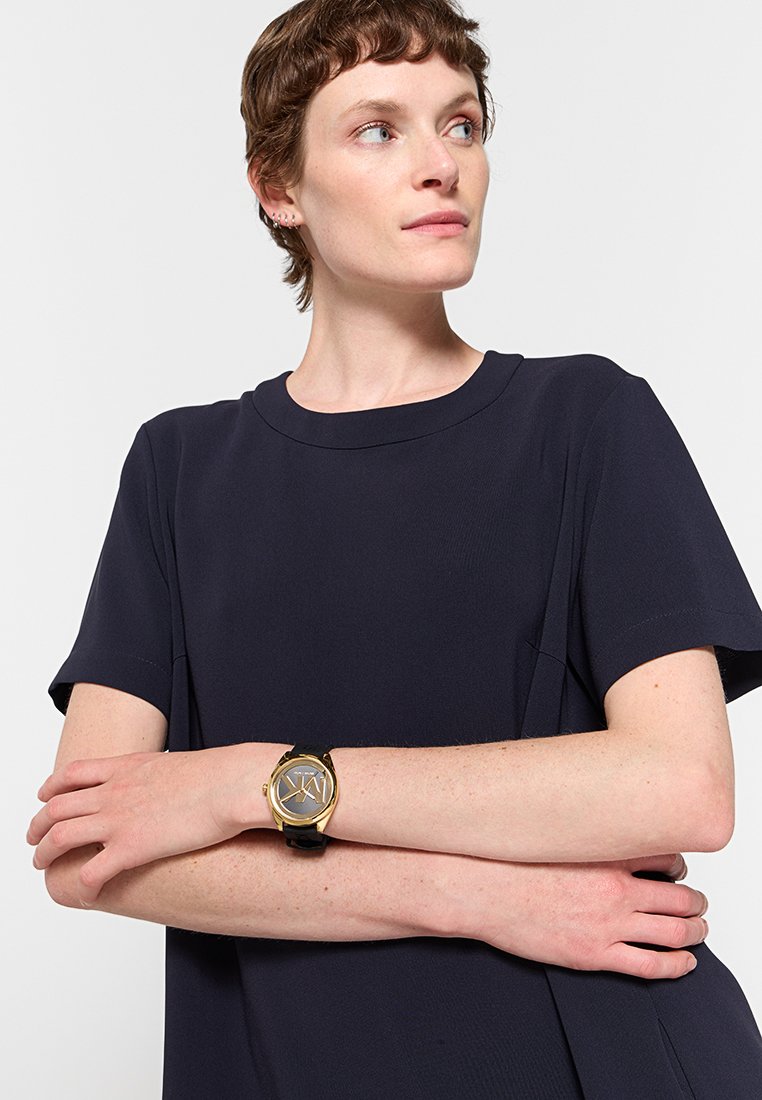 Camisa negra de manga corta con cuello redondo, combinada con un reloj acentuado en dorado que presenta una esfera circular y una correa negra.