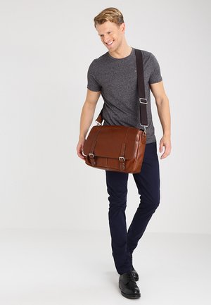 Sac messager en cuir marron avec une bandoulière réglable, quincaillerie en métal, deux boucles à l'avant et une texture lisse, tenu par une personne en tenue décontractée.