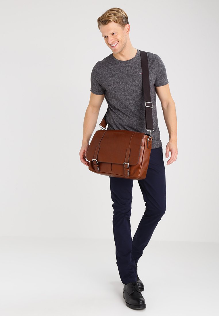 Sac messager en cuir marron avec une bandoulière réglable, quincaillerie en métal, deux boucles à l'avant et une texture lisse, tenu par une personne en tenue décontractée.