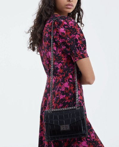 Borsa nera in pelle effetto coccodrillo con tracolla in catena argentata, forma rettangolare e chiusura a pattina. Indossata sopra un vestito con motivo floreale.