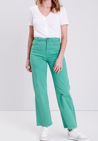 Pantalones verdes de talle alto con un diseño de pierna recta y dos bolsillos frontales. Combinados con una blusa blanca de botonadura y zapatillas blancas.