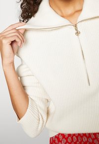 Gerippter cremefarbener Pullover mit hohem Kragen und goldfarbenem Reißverschluss. Weiche Textur, lockere Passform, getragen mit einem gemusterten roten Rock.