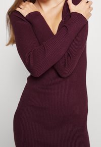 Mujer vestida con un vestido de punto acanalado de manga larga y ajustado en burdeos profundo, cruzando los brazos sobre el pecho.