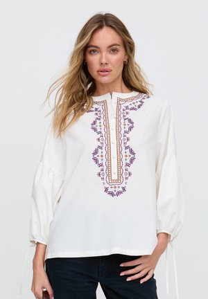 Frau mit langen Haaren, die eine weiße Bluse mit lila und brauner Stickerei und Knopfleiste trägt, steht vor einem einfarbigen hellen Hintergrund.